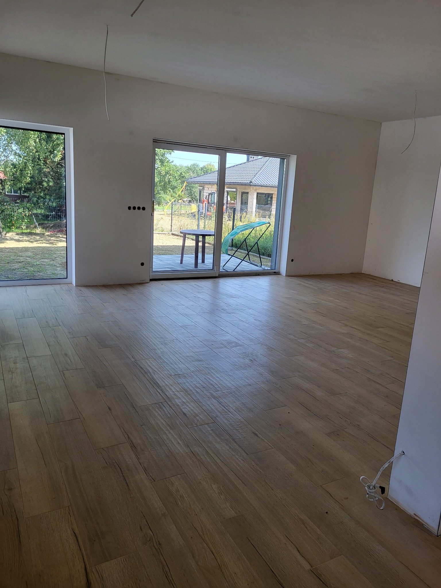 Wnętrze w stanie deweloperskim w Orzeszu z ułożonymi panelami podłogowymi imitującymi drewno, widoczne okna balkonowe i wystające kable ze ściany.