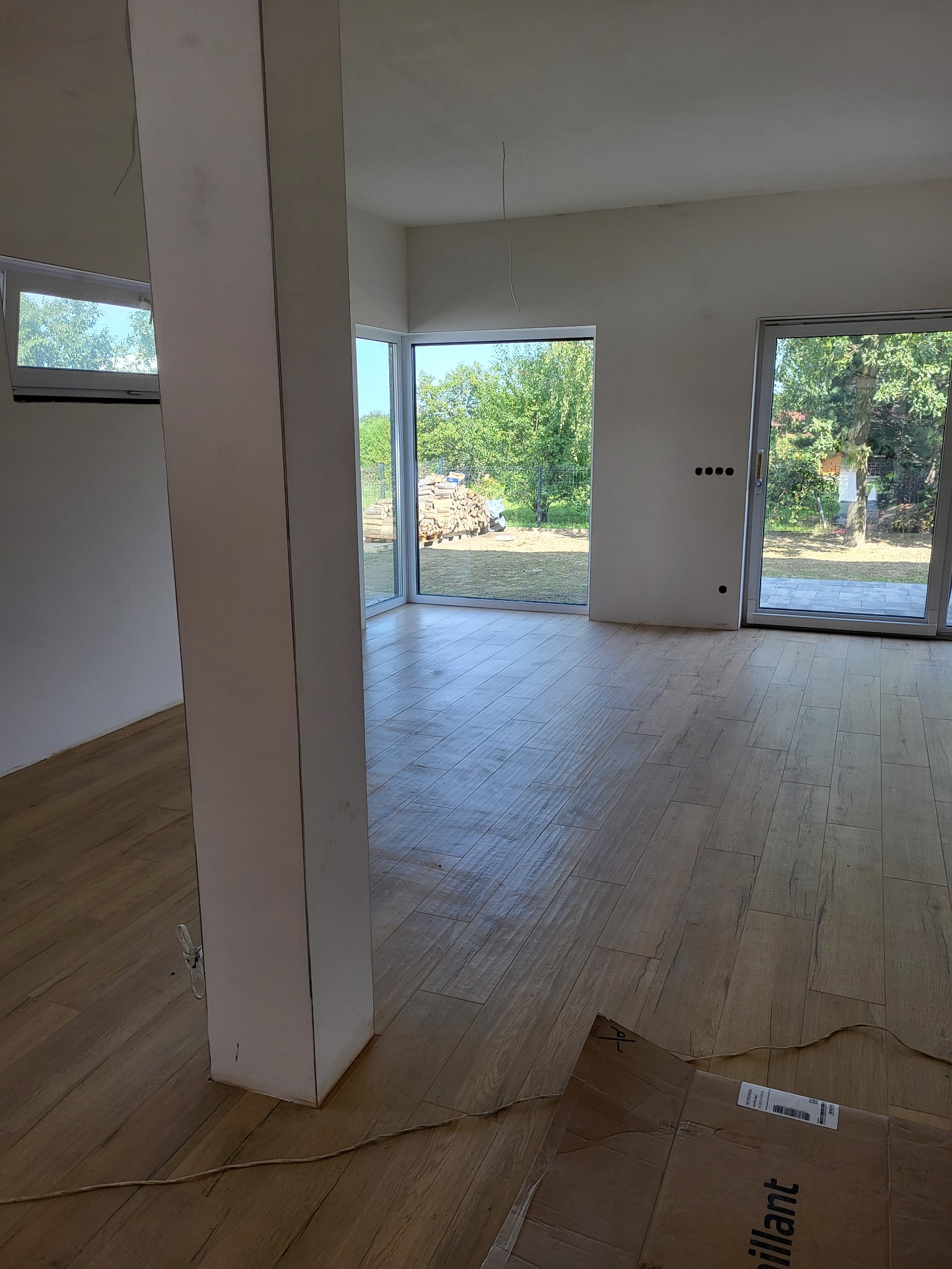 Jasne wnętrze z drewnopodobną podłogą, widoczne okna z widokiem na ogród, karton na podłodze, kolumna w centrum pokoju. Częściowo wykończone ściany.