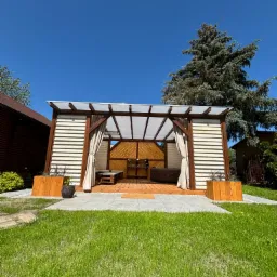 Nowoczesna pergola z drewnianą konstrukcją, zasłonami i drewnianym tarasem w Gniewkowie, z widocznym jacuzzi wewnątrz, otoczona zielonym trawnikiem i drzewami.