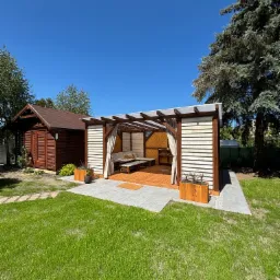 Nowoczesna, drewniana konstrukcja z zasłonami w ogrodzie, obok drewniany domek narzędziowy, na trawniku drewniane donice z roślinami.