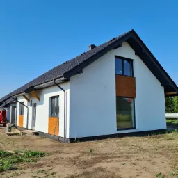 Nowoczesny, parterowy dom z białymi ścianami, czarnym dachem i drewnianymi akcentami elewacyjnymi; widoczne przygotowane podłoże pod trawnik.