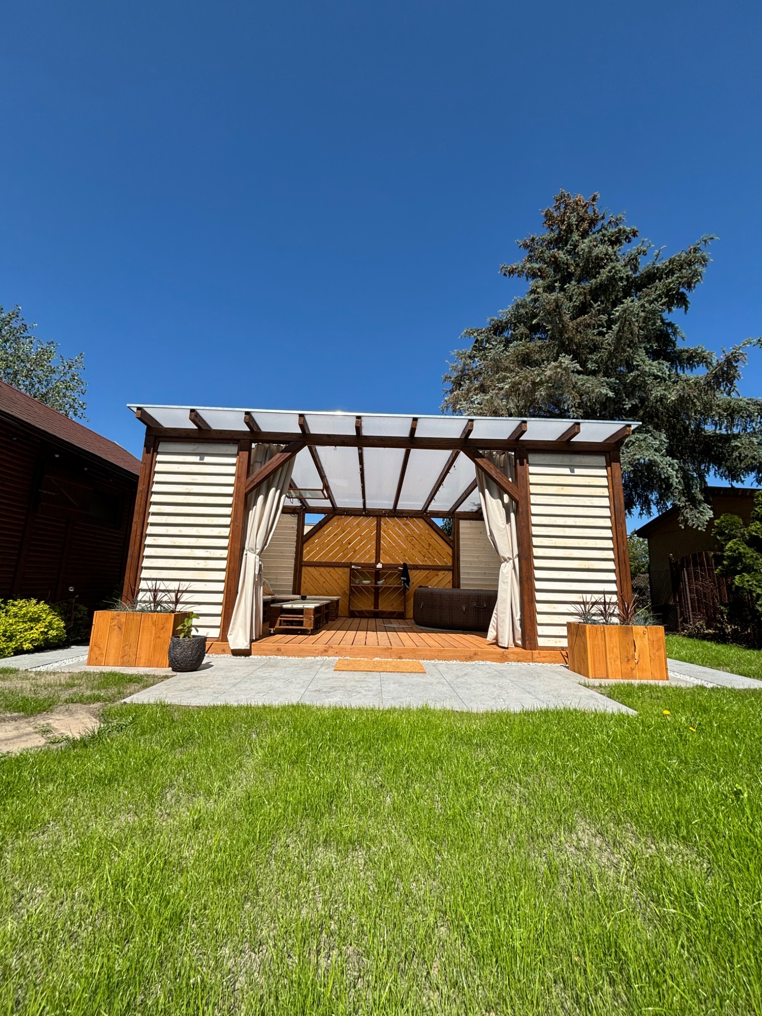 Nowoczesna pergola z drewnianą konstrukcją, zasłonami i drewnianym tarasem w Gniewkowie, z widocznym jacuzzi wewnątrz, otoczona zielonym trawnikiem i drzewami.
