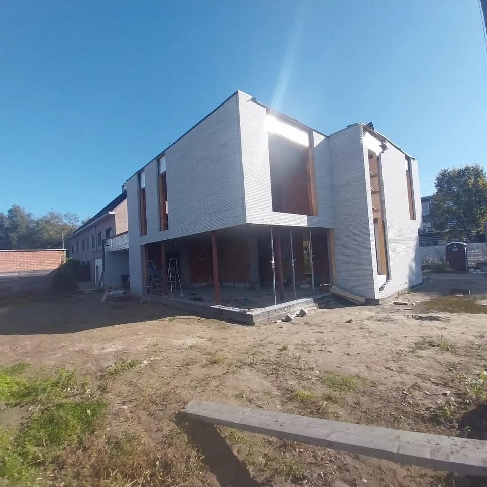 Nowoczesny dom w trakcie budowy, z widoczną konstrukcją nośną, elewacją z szarej cegły i dużymi oknami bez szyb, na placu budowy z widocznymi narzędziami i toaletą przenośną w tle.