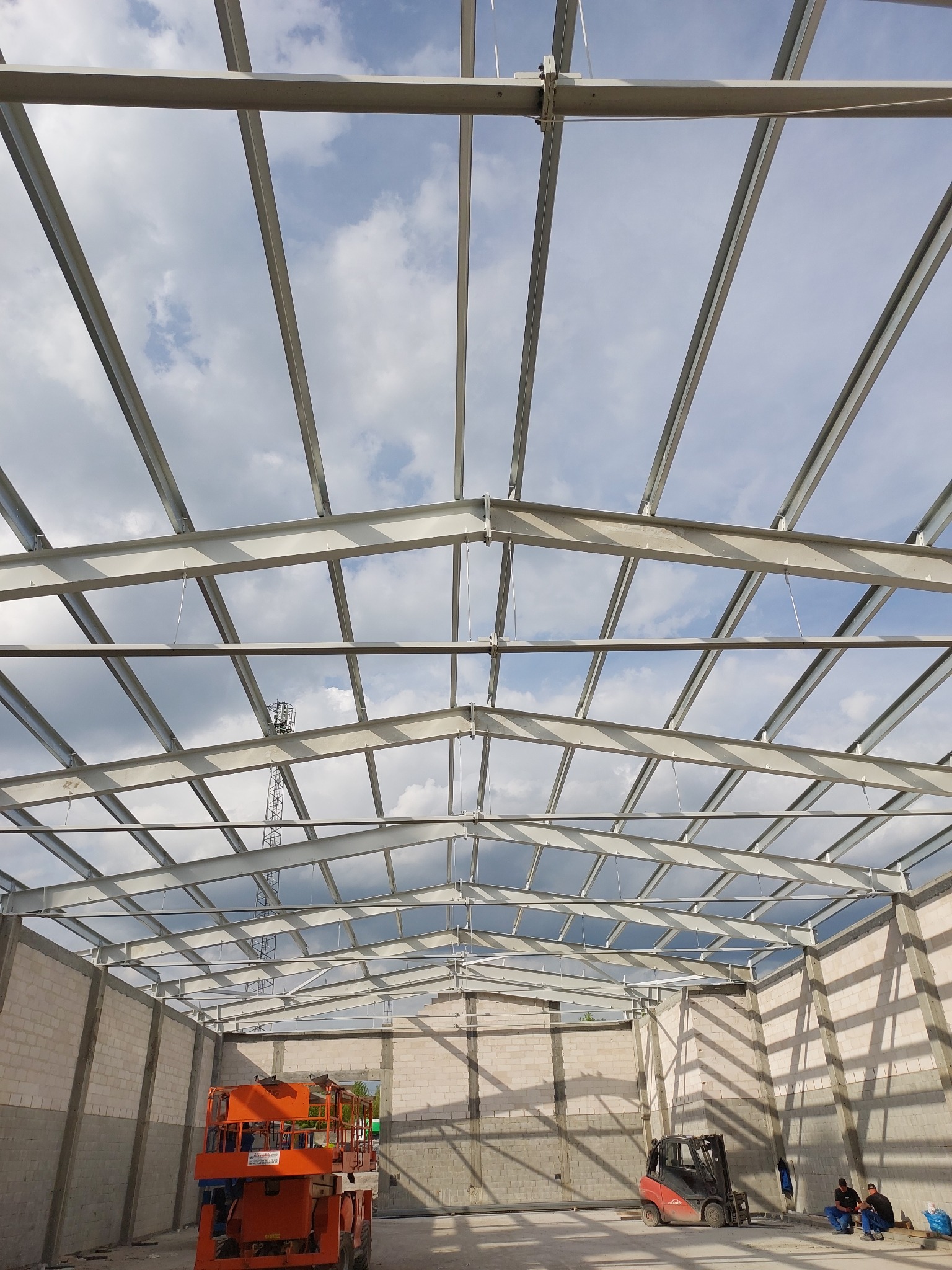 Wnętrze budowanej hali z widoczną konstrukcją stalową dachu na tle pochmurnego nieba, z pomarańczowym podnośnikiem i czerwonym wózkiem widłowym wewnątrz, oraz dwoma osobami siedzącymi obok wózka.