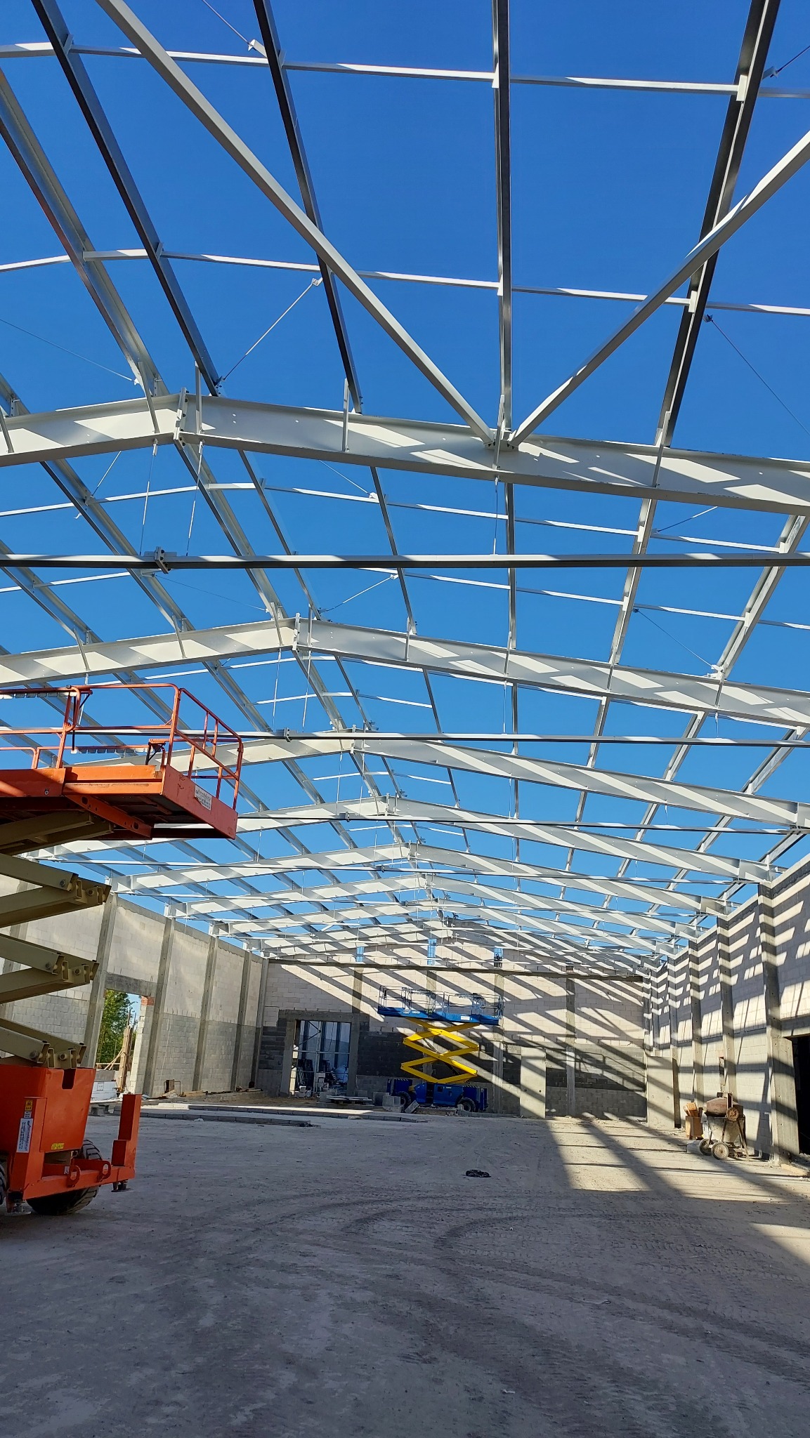 Wnętrze powstającej hali z widoczną konstrukcją stalową dachu i ścian. Na pierwszym planie pomarańczowy podnośnik nożycowy, w tle drugi, żółty podnośnik. Wyraźne światło słoneczne tworzy długie...