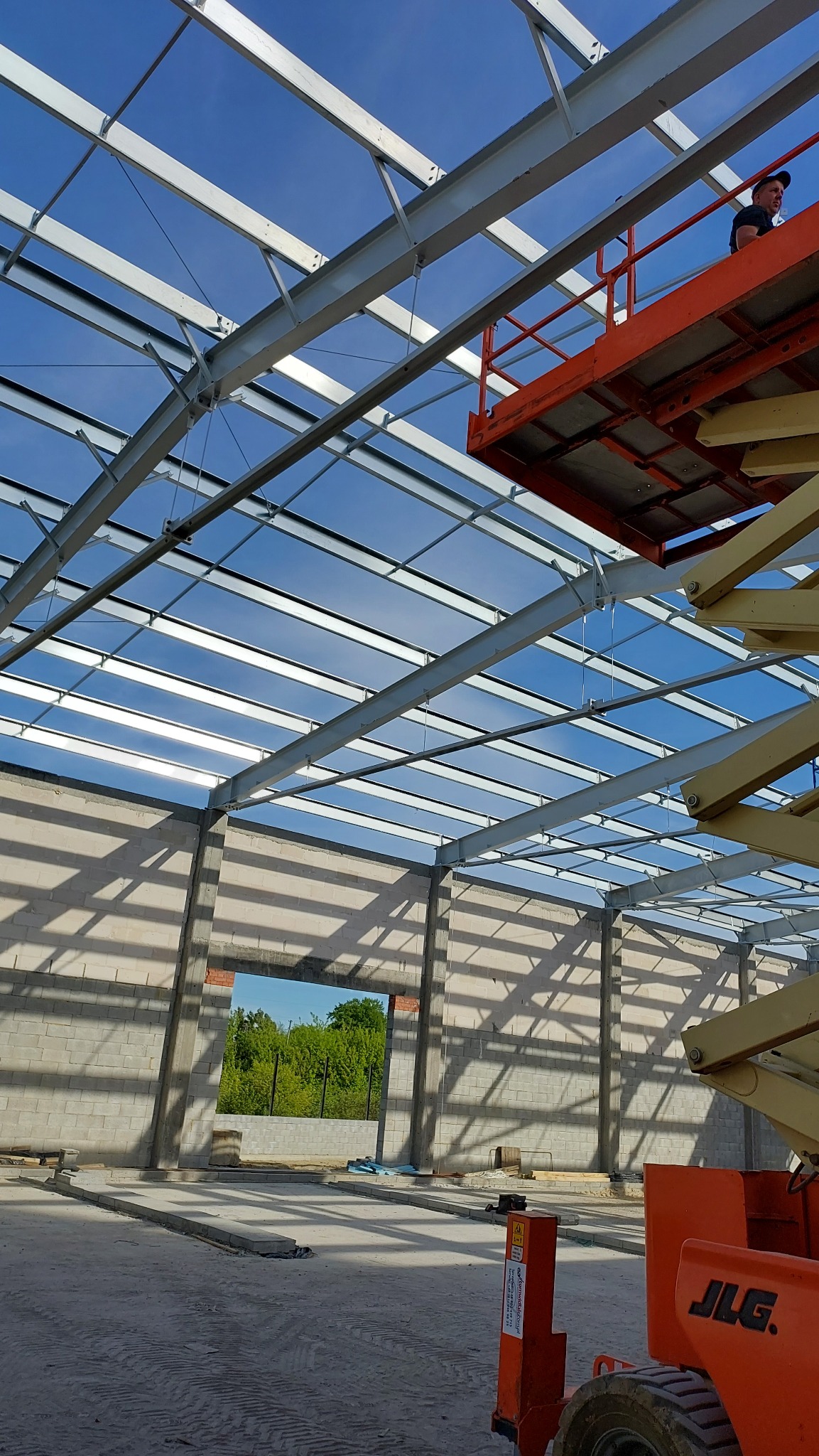 Wnętrze budowanej hali z widoczną konstrukcją stalową dachu i fragmentem ściany z betonu komórkowego; na pierwszym planie pomarańczowy podnośnik koszowy z pracownikiem na górze.
