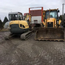 Żółty minikoparka Neuson i koparko-ładowarka JCB w Nowym Gronowie, stojące na ubitym, błotnistym gruncie, z widocznymi śladami gąsienic i opon.