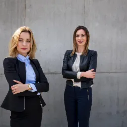 Dwie kobiety w formalnych strojach stojące na tle szarej betonowej ściany z rękami skrzyżowanymi na piersiach, prezentujące profesjonalny wizerunek.