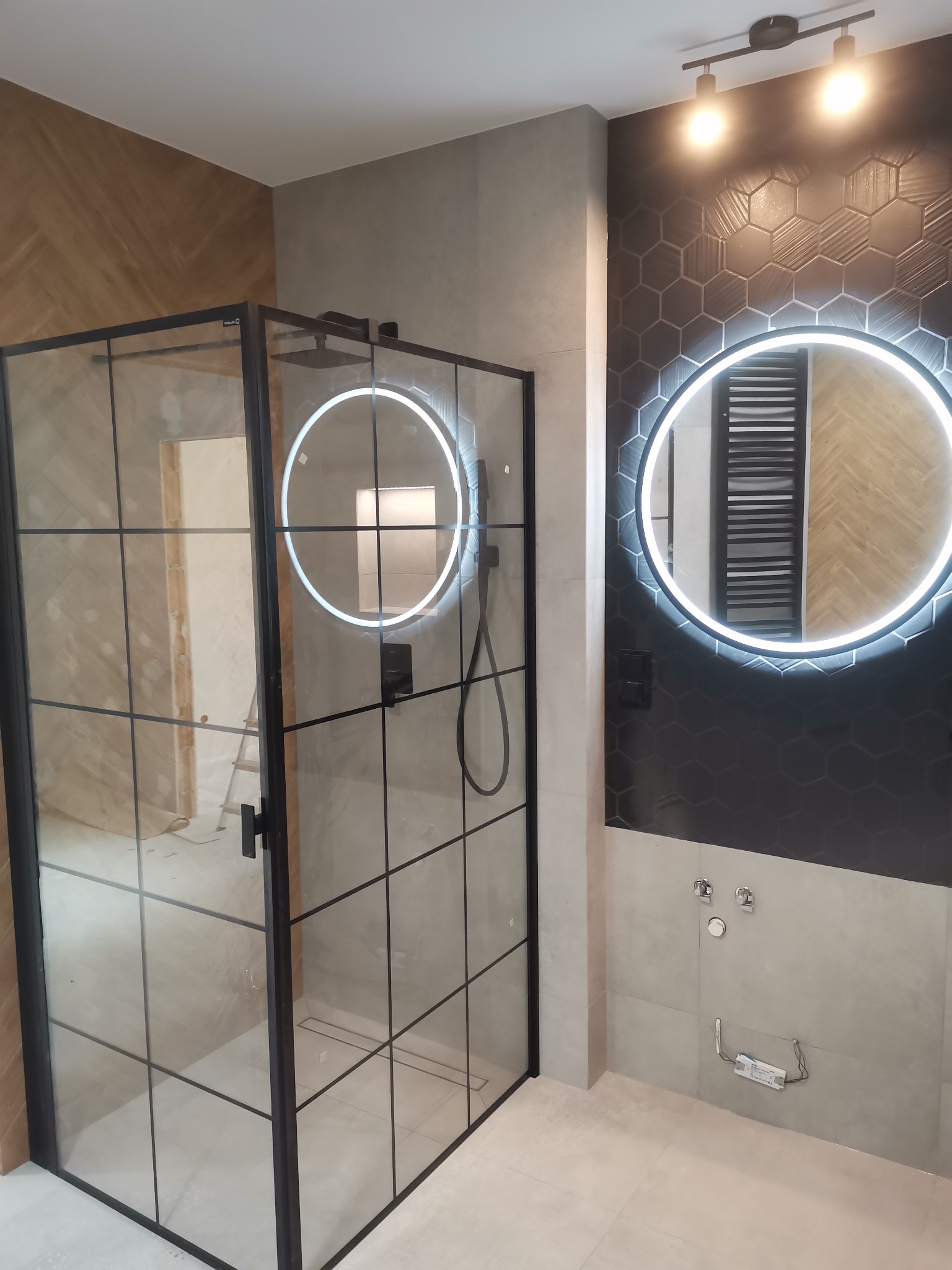 Nowoczesna łazienka z kabiną prysznicową typu walk-in z czarnymi szprosami, okrągłym lustrem z podświetleniem LED i heksagonalnymi płytkami na ścianie.