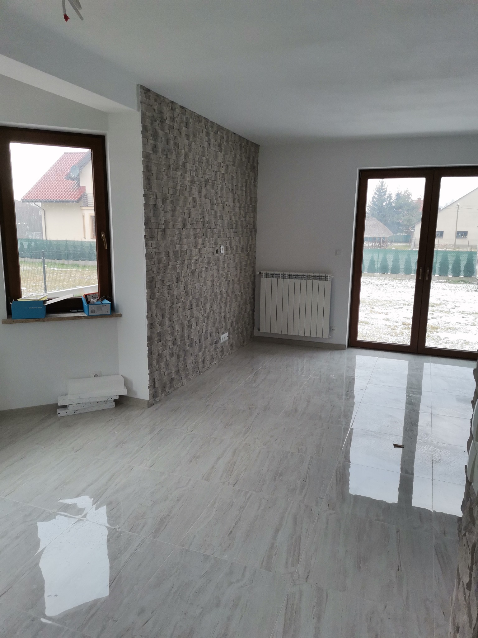 Wykończone wnętrze z szarą, kamienną okładziną na ścianie, jasnymi płytkami na podłodze z połyskiem, białym grzejnikiem i oknami z widokiem na zimowy krajobraz.