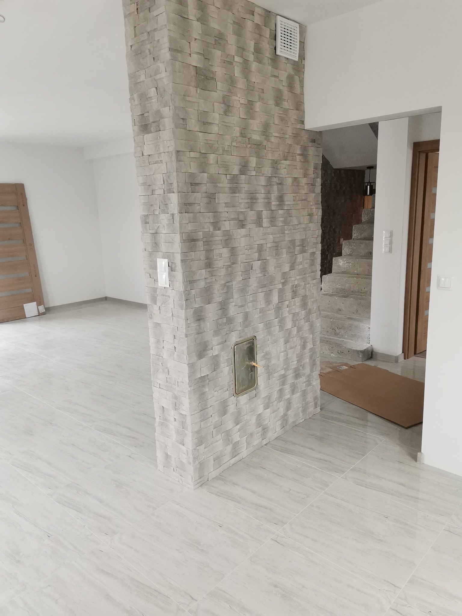 Świeżo wykończone wnętrze z kolumną obłożoną dekoracyjną, szarą okładziną kamienną, pasującą do marmurowej podłogi, widoczny otwór rewizyjny w kolumnie oraz częściowo widoczne schody w tle.