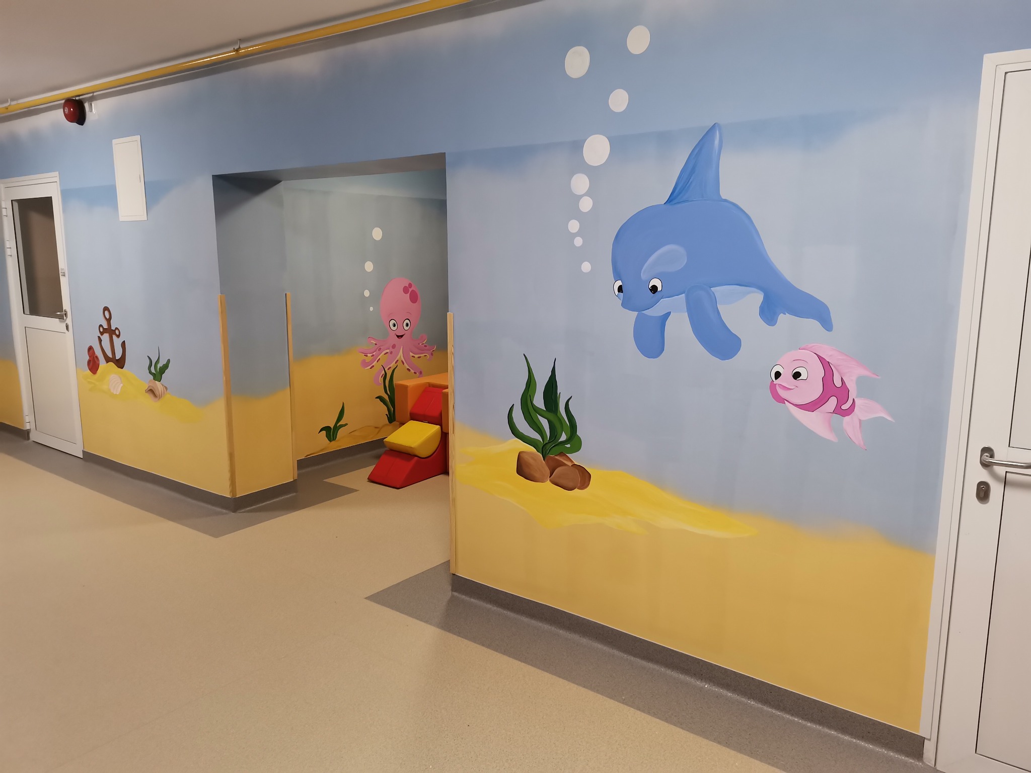 Korytarz w placówce medycznej z dekoracyjnym malowaniem ścian przedstawiającym podwodny świat: delfin, ryba, ośmiornica, kotwica, algi i bąbelki powietrza.