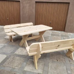 Surowy drewniany zestaw ogrodowy: stół i dwie ławki z oparciami, na tle garażu z brązowymi drzwiami segmentowymi.