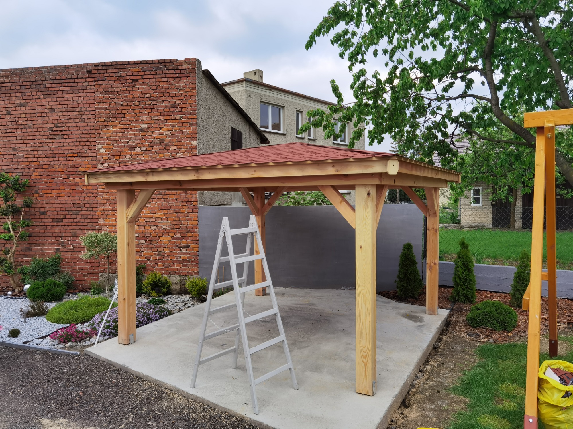 Nowo wybudowana drewniana altana z czerwoną dachówką na betonowej podstawie, z drabiną opartą o jedną z kolumn, w tle budynek z cegły i zielony ogród.
