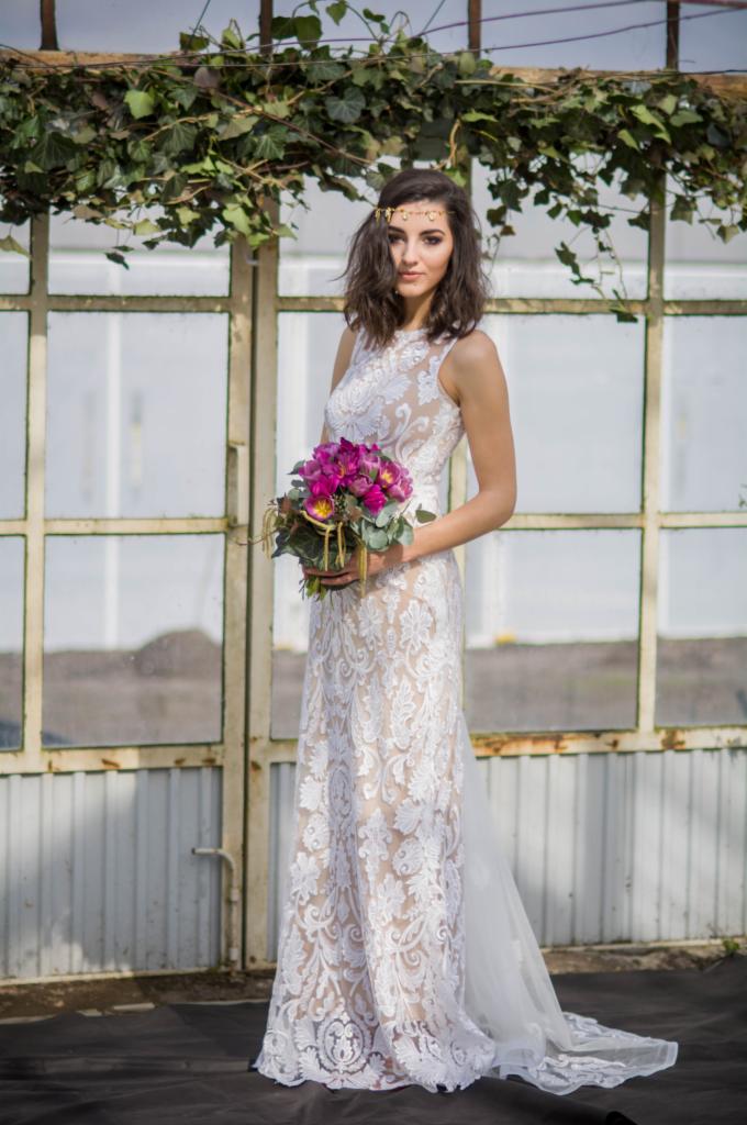 Panna młoda w koronkowej sukni ślubnej z bukietem fioletowych tulipanów, pozuje na tle szklarni porośniętej bluszczem.