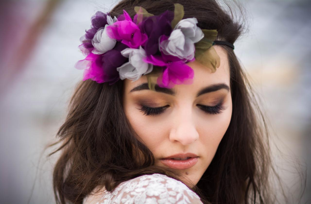 Portret kobiety z ciemnymi włosami, zamkniętymi oczami i kwiatowym wiankiem w odcieniach fioletu i szarości na głowie, delikatny makijaż i biała koronkowa bluzka.