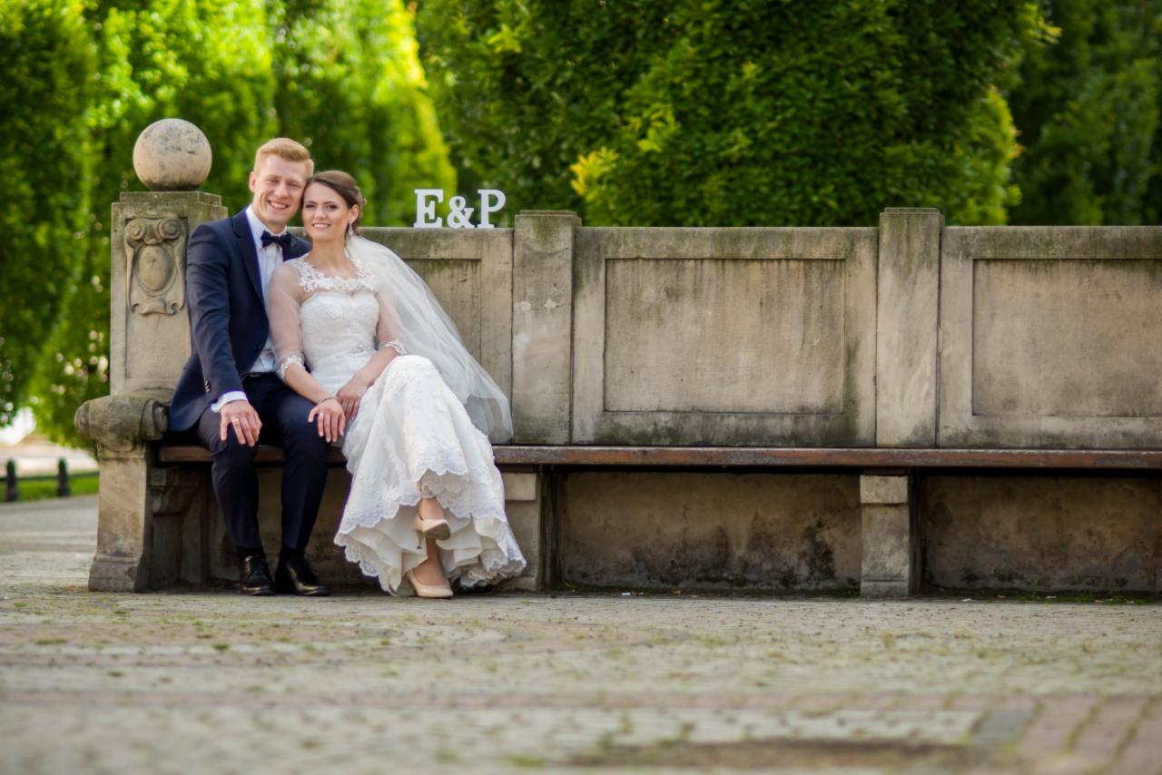 Uśmiechnięta para młoda siedzi na kamiennej ławce w parku, za nimi napis E&P z białych liter.