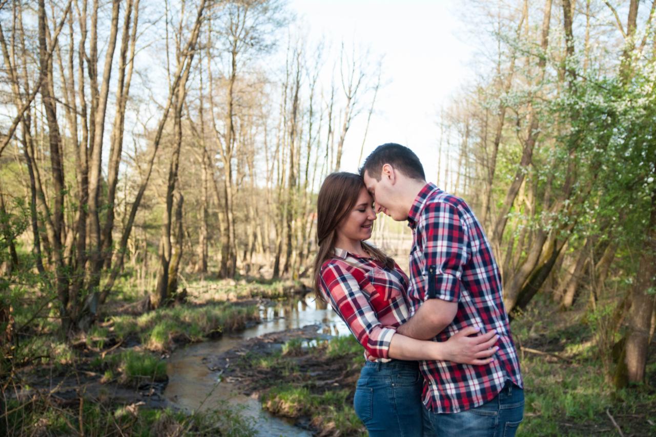 Zakochana para w czerwono-białych koszulach w kratę, obejmująca się na tle leśnego strumienia i drzew bez liści.