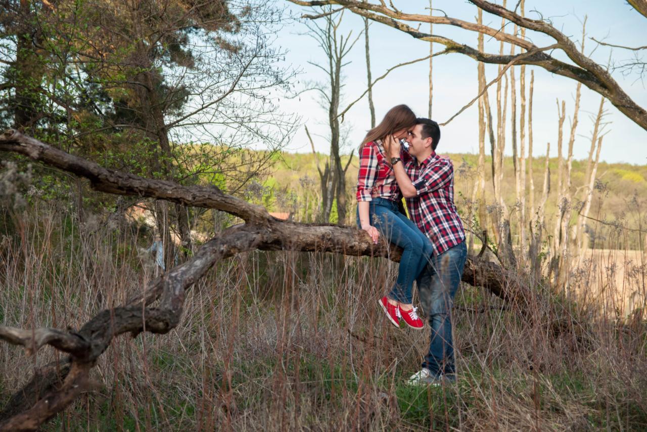 Para w czerwonych koszulach w kratę przytula się na gałęzi drzewa w zaroślach, ona siedzi, on stoi obok.