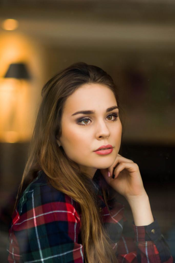 Portret kobiety w kraciastej koszuli z ręką pod brodą, wykonany przez szybę z widocznym odbiciem lampy w tle.