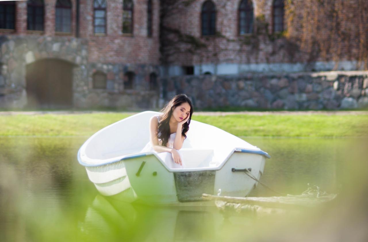 Kobieta w jasnej sukience siedzi w łódce na wodzie, z zamyślonym wyrazem twarzy, na tle kamiennego budynku z cegły porośniętego bluszczem.