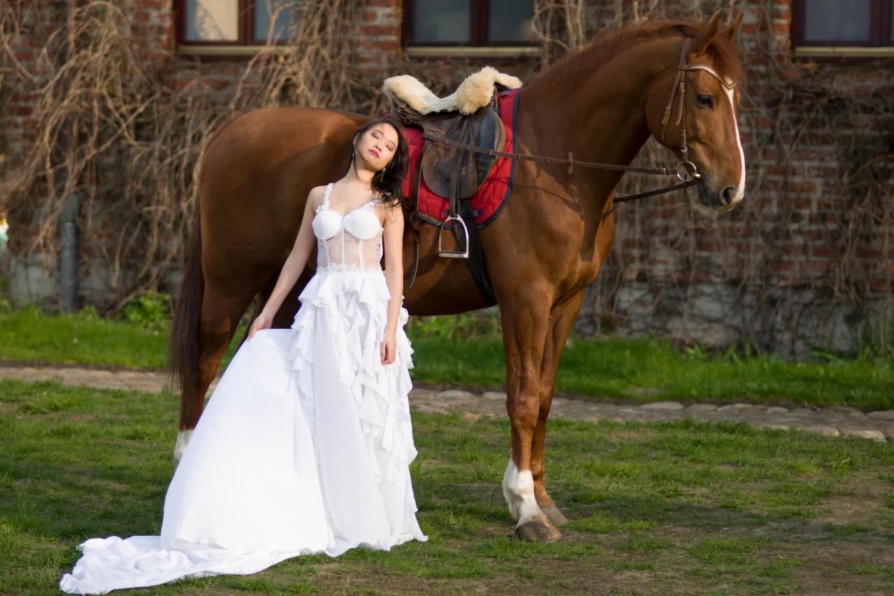 Kobieta w białej sukni pozuje opierając się o brązowego konia z siodłem na tle ceglanego budynku porośniętego bluszczem.