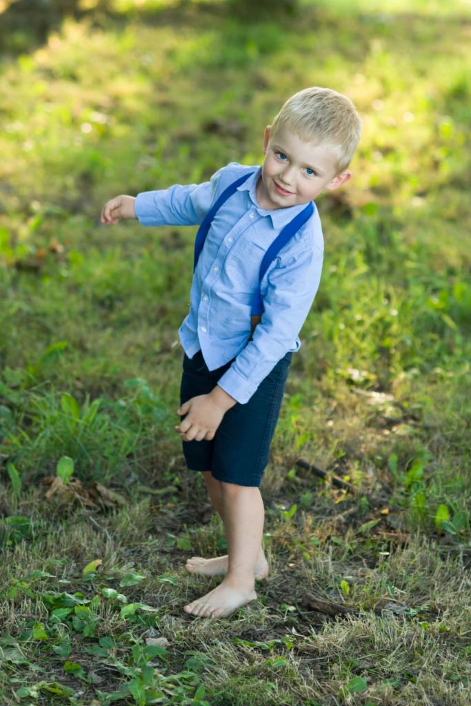 Uśmiechnięty, boso stojący chłopiec w błękitnej koszuli i granatowych szortach z szelkami pozuje na trawie w słoneczny dzień.