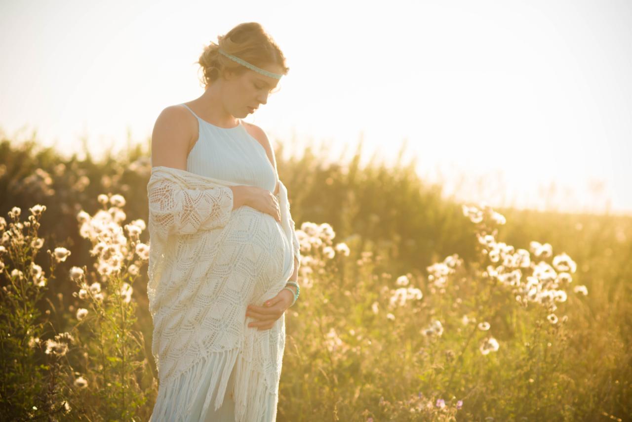 Kobieta w ciąży w jasnej sukience i ażurowym szalu pozuje w polu kwitnących roślin, z delikatnym oświetleniem zachodzącego słońca i turkusową opaską na głowie.