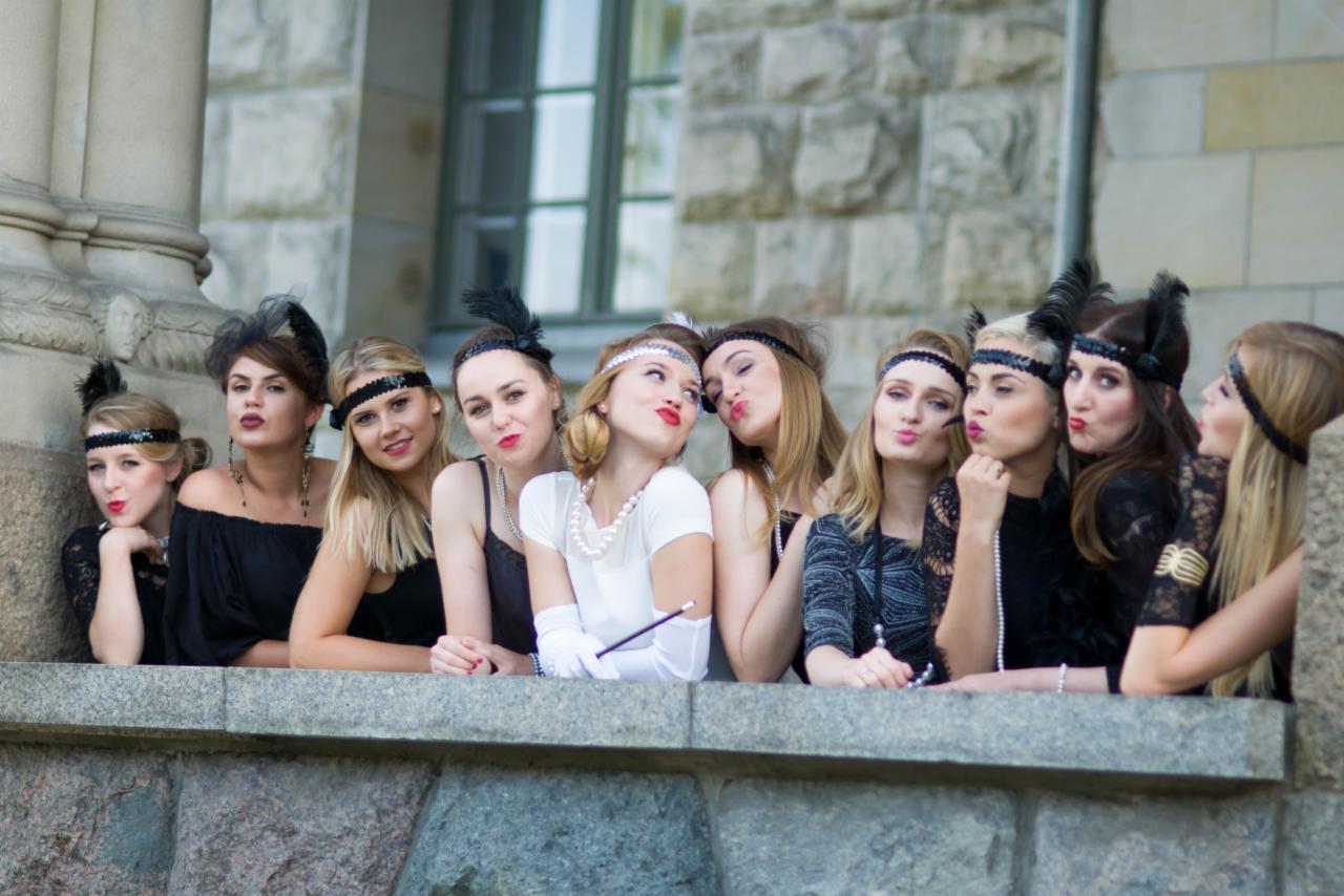 Grupa kobiet w stylizacjach z lat 20. XX wieku, z piórami we włosach i perłami, pozuje na tle kamiennej fasady budynku.