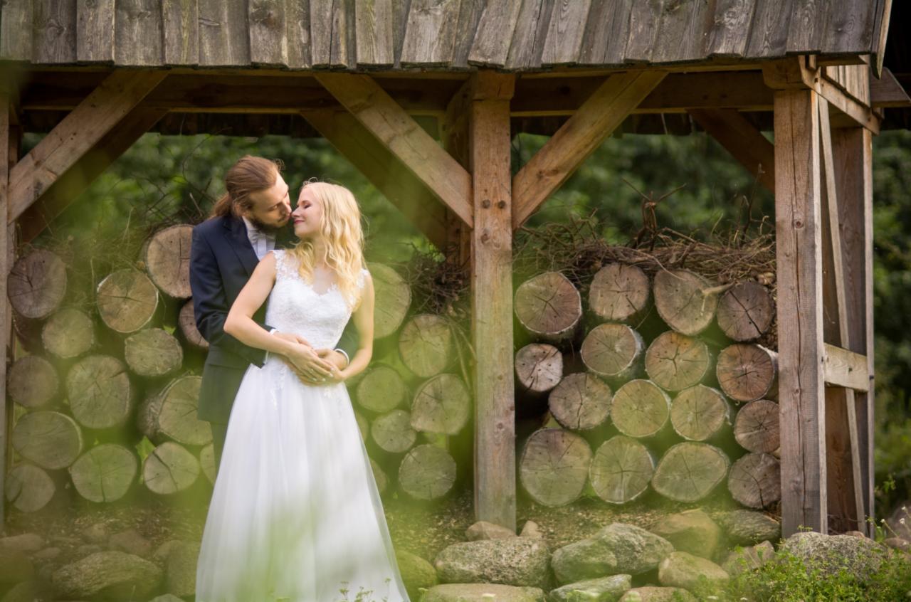 Para młoda w objęciach pod drewnianą konstrukcją z ułożonymi w tle pniakami drzew, delikatne światło, naturalna sceneria.