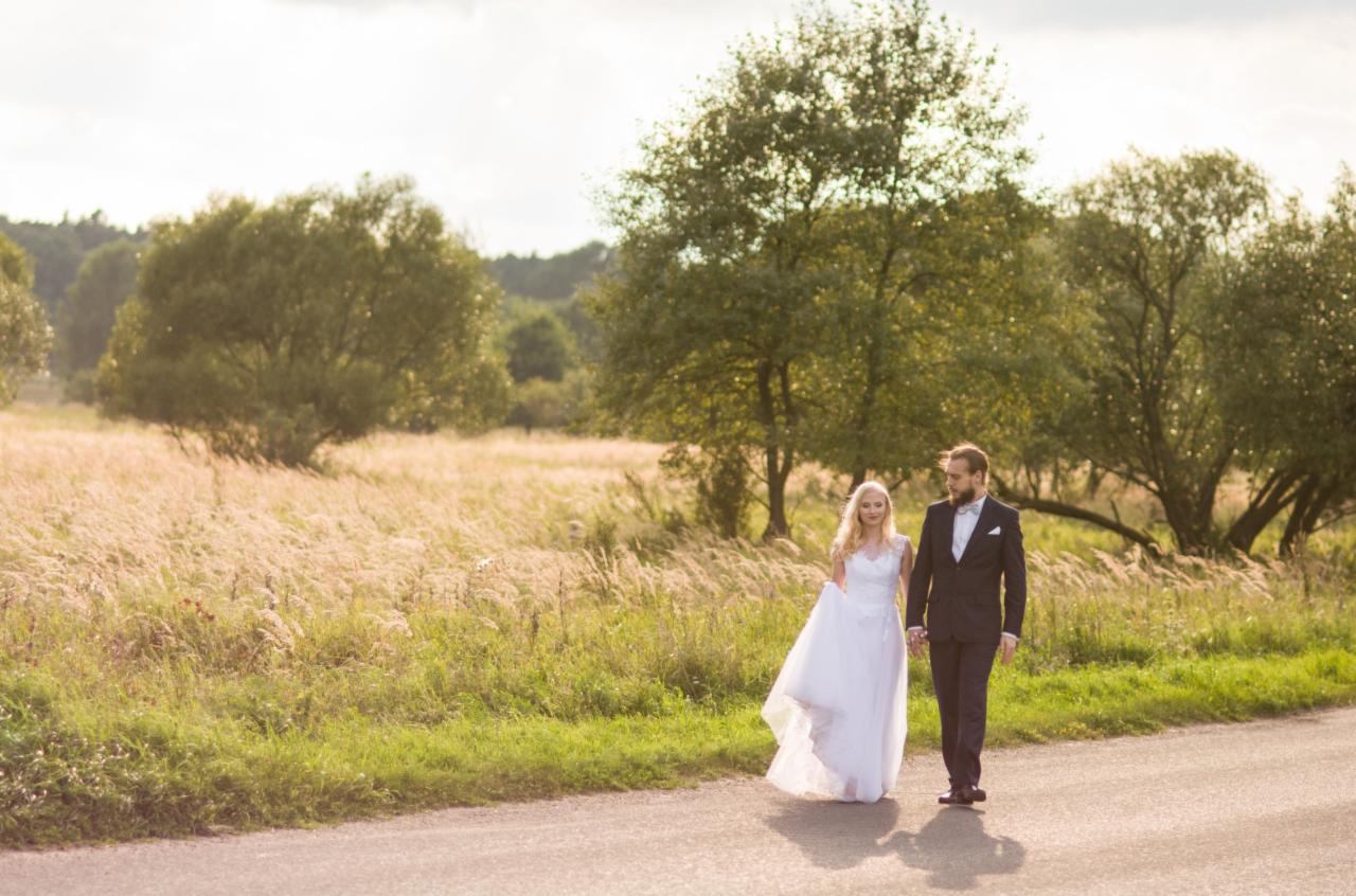 Para młoda spaceruje polną drogą w słoneczny dzień, wysoka trawa i drzewa w tle.