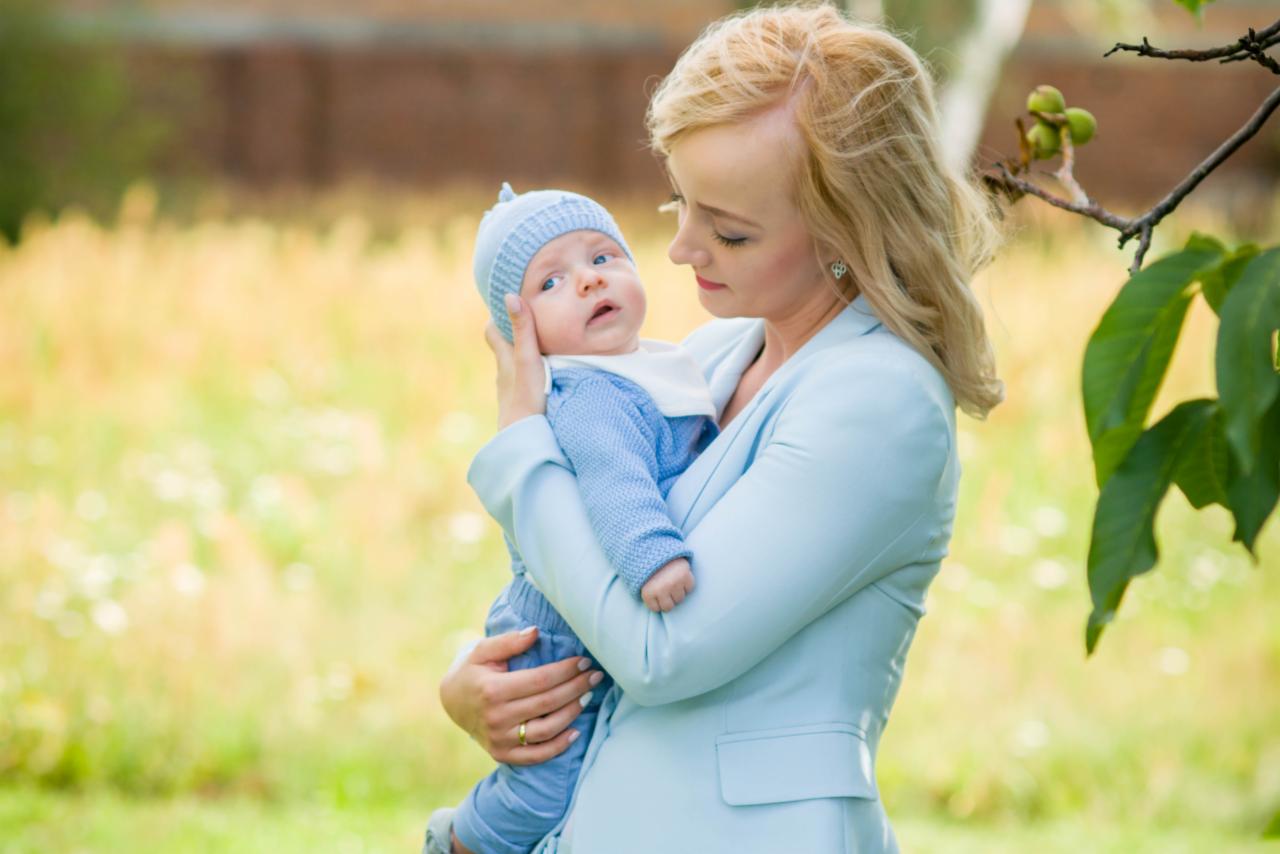 Matka w pastelowo-błękitnym żakiecie trzyma na rękach niemowlę w błękitnym ubranku i czapeczce, z zielonym tłem łąki i gałęzi drzewa z liśćmi.