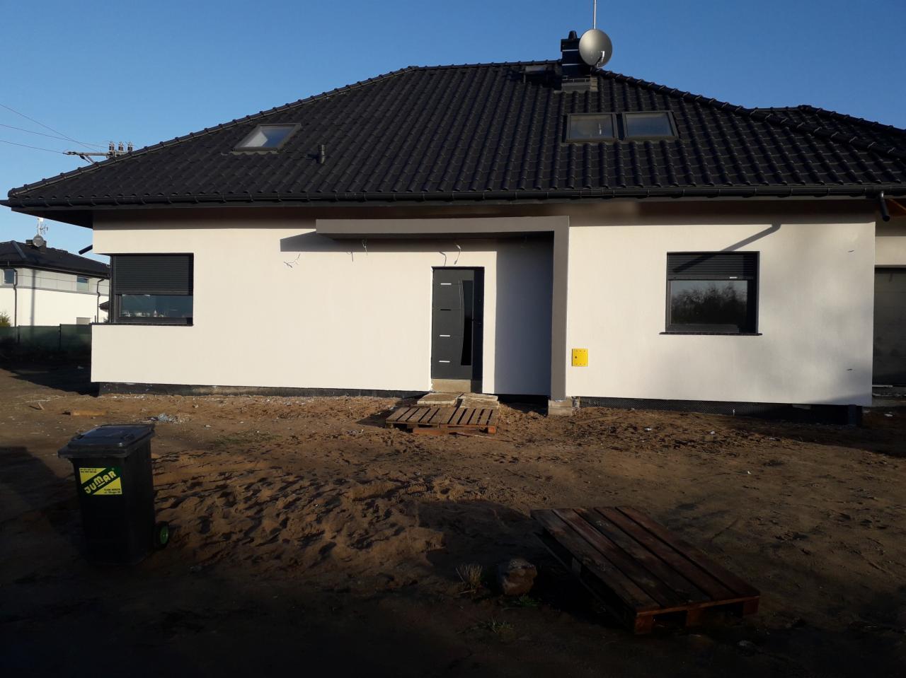 Świeżo otynkowany dom jednorodzinny z czarną dachówką i ramami okiennymi, widoczne palety przed wejściem i pojemnik na śmieci po lewej stronie.