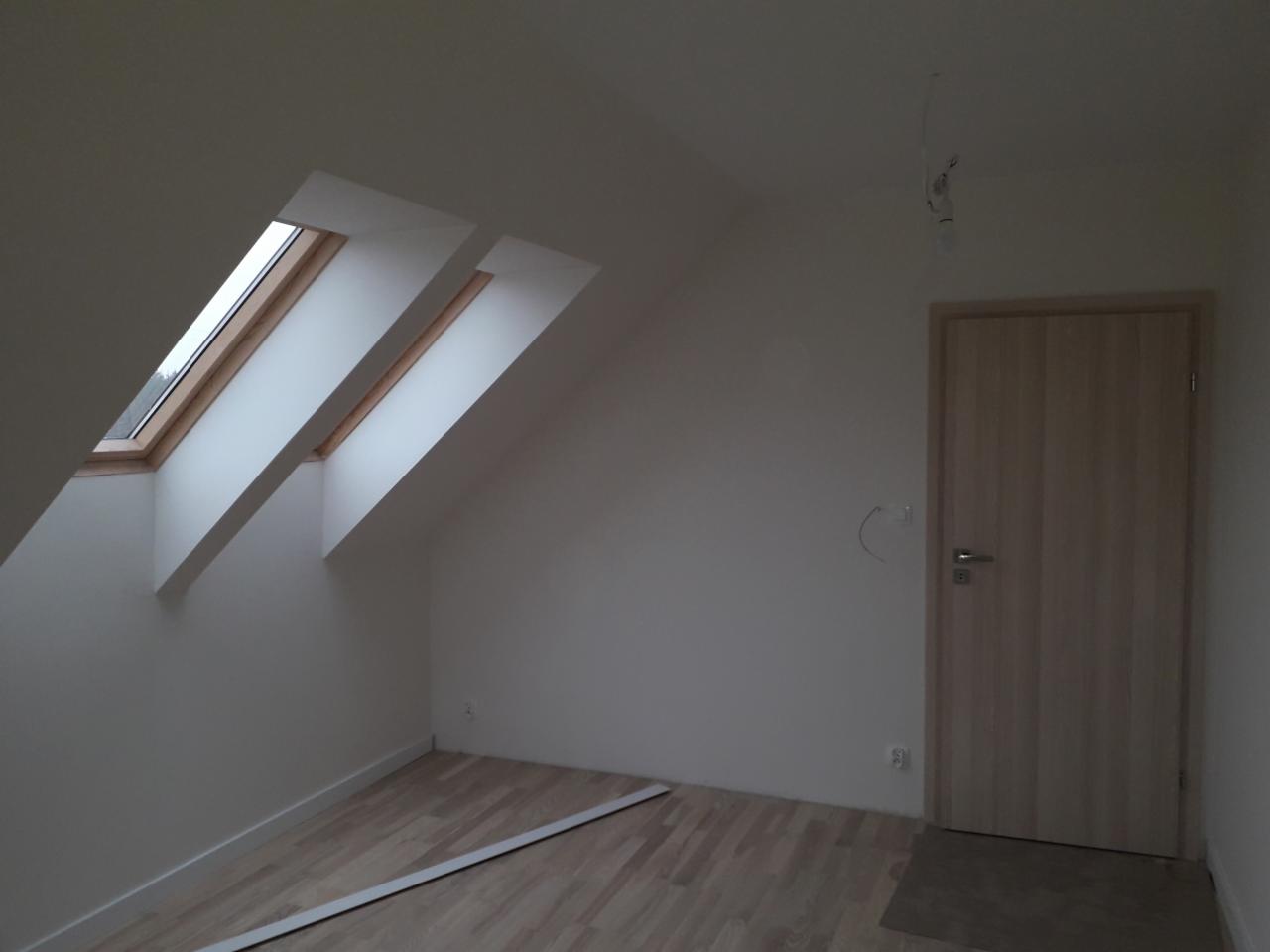 Poddasze po remoncie z dwoma oknami dachowymi, świeżo pomalowanymi ścianami na biało, drewnianą podłogą i jasnymi drzwiami. Widoczne niedociągnięcia w postaci wystających kabli i niedokończonej...