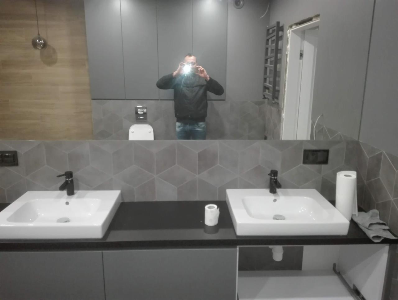 Nowoczesna łazienka z dwoma białymi umywalkami nablatowymi na szarym blacie, szare płytki w geometryczny wzór na ścianie, widoczne odbicie osoby robiącej zdjęcie w dużym lustrze.