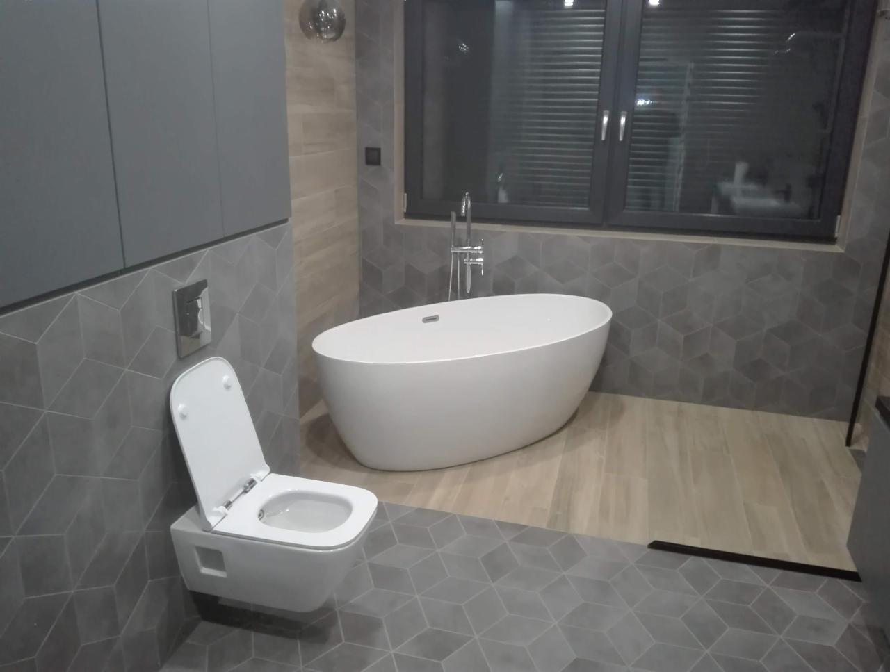 Nowoczesna łazienka z wolnostojącą wanną, podwieszaną toaletą z otwartą deską, szarą geometryczną glazurą i drewnopodobną podłogą w strefie prysznica.