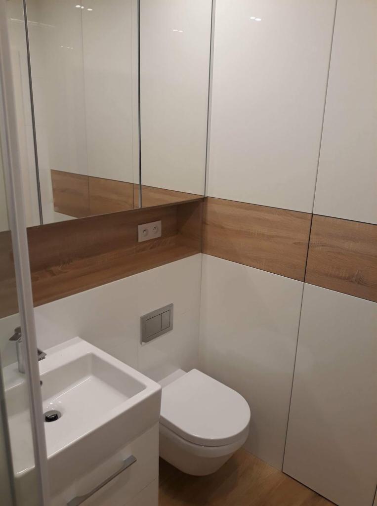 Nowoczesna łazienka z białą ceramiką, podwieszaną miską WC, umywalką z szafką, lustrem i drewnianymi akcentami na ścianach.