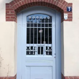 Odnowione wejście do budynku z łukowym portalem z czerwonej cegły, pomalowane na jasnoniebiesko drzwi z ozdobnymi kratami i kwiatowymi motywami, tabliczka z numerem 5 i oznaczenie zabytku.