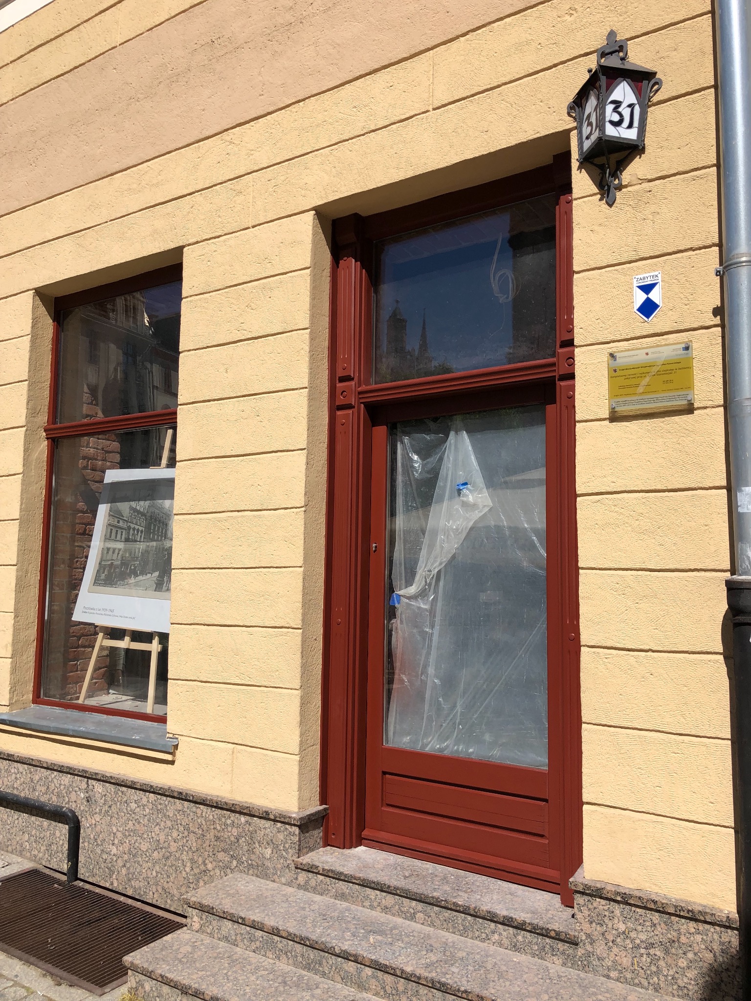 Renowacja stolarki okiennej i drzwiowej zabytkowej kamienicy na starym rynku w Toruniu, okna zabezpieczone folią malarską, widoczna tabliczka informacyjna konserwatora zabytków oraz numer budynku 31.