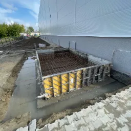 Przygotowanie fundamentu pod budynek przemysłowy: szalunek z żółtymi wkładkami Doka, zbrojenie stalowe wewnątrz, teren wokół zalany wodą.