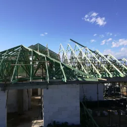 Wznoszenie konstrukcji dachu z impregnowanego drewna na murowanym domu z białych bloczków, widok na niebo z lekkimi chmurami.