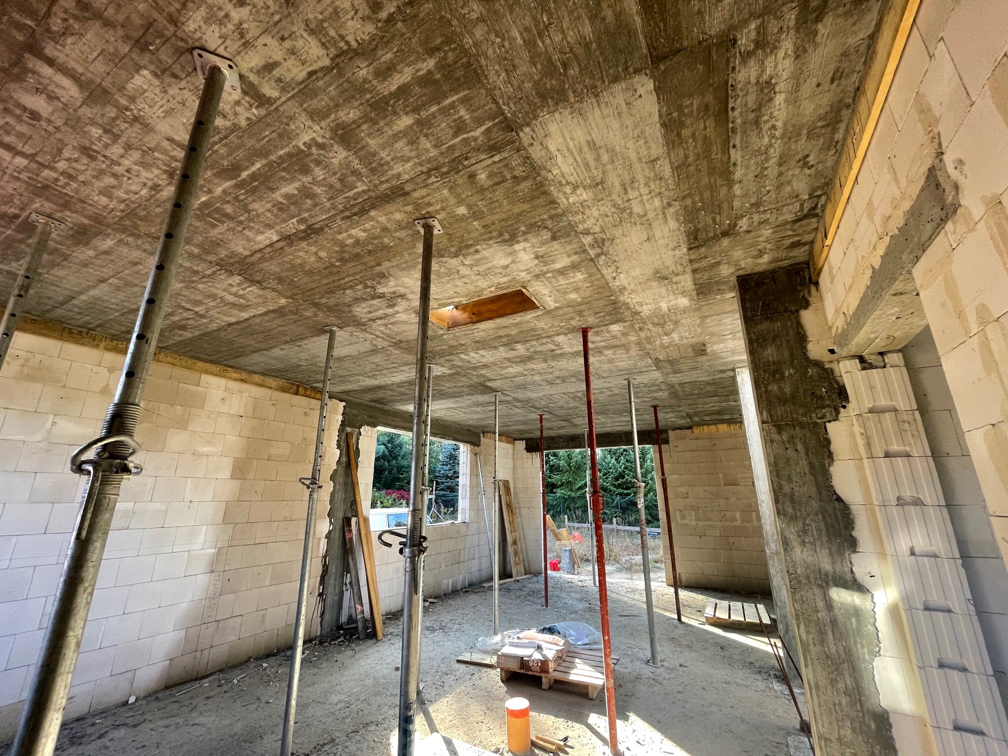 Wnętrze budynku w trakcie budowy z widocznym betonowym stropem wspartym na stalowych podporach, ściany z bloczków, otwory okienne i drzwiowe, oraz paleta z materiałami budowlanymi na podłodze.
