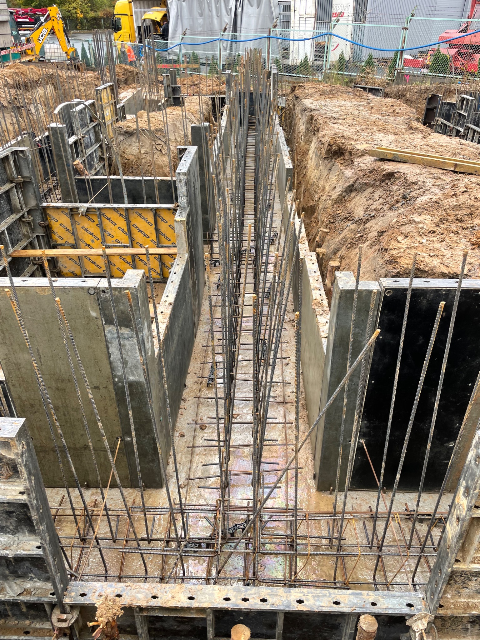 Widok z góry na wykop z przygotowanymi szalunkami i zbrojeniem pod fundament. Żółty dźwig JCB w tle. Widoczne elementy szalunków Doka.