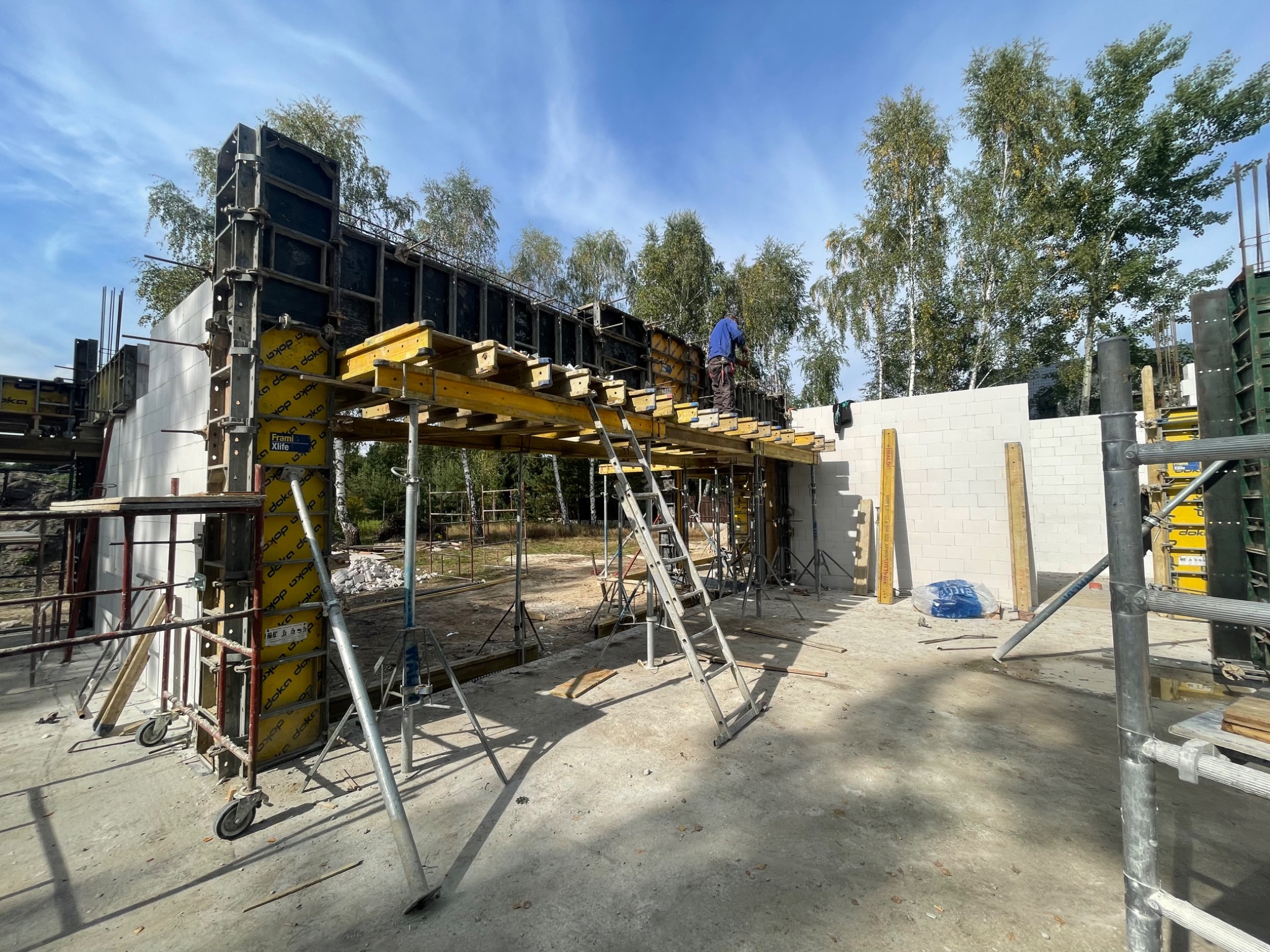 Konstrukcja szalunkowa z żółtymi podporami na budowie domu, widoczny pracownik w niebieskiej koszuli na górze konstrukcji, ściany z bloczków betonu komórkowego w tle, drzewa i niebieskie niebo