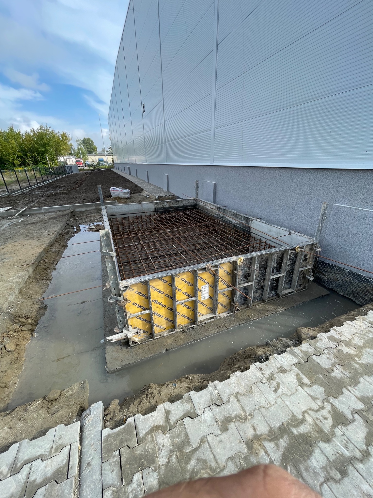 Przygotowanie fundamentu pod budynek przemysłowy: szalunek z żółtymi wkładkami Doka, zbrojenie stalowe wewnątrz, teren wokół zalany wodą.