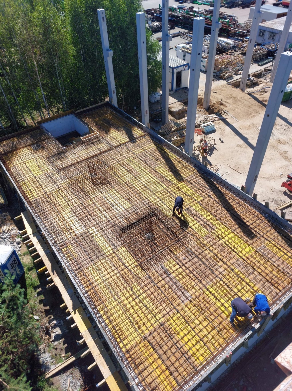 Widok z góry na zbrojenie stropu budynku w trakcie budowy, z widocznymi pracownikami w kombinezonach, wystającymi słupami konstrukcyjnymi i żółtą membraną izolacyjną pod zbrojeniem.