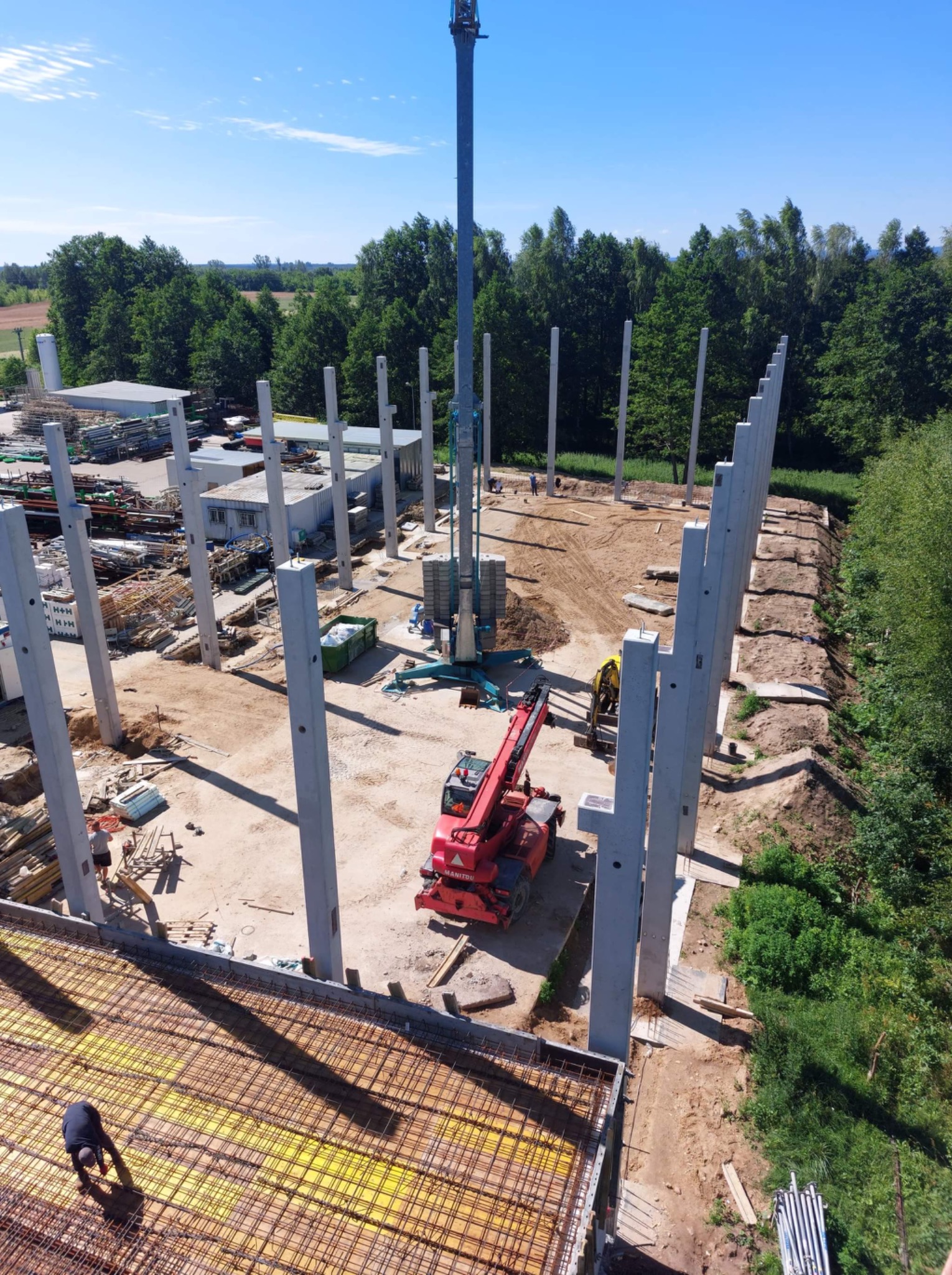 Wysoki kąt widzenia na plac budowy z prefabrykowanymi słupami żelbetowymi, dźwigiem teleskopowym Manitou i zbrojeniem płyty fundamentowej z żółtym podkładem, otoczony zielenią.