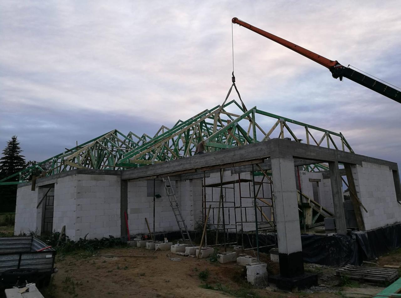 Wznoszenie konstrukcji dachu z drewnianych wiązarów na budynku w stanie surowym otwartym, z użyciem pomarańczowego dźwigu, widok na szkielet domu z betonu komórkowego i betonowe nadproża.
