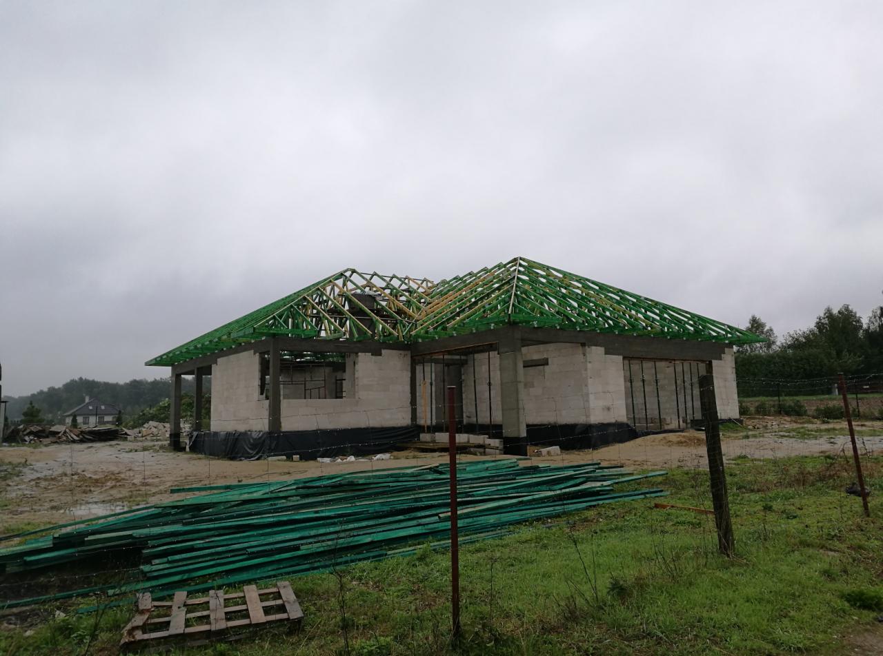Niedokończony stan surowy otwarty domu z betonu komórkowego z zieloną konstrukcją dachu, widoczne deski na pierwszym planie i pochmurne niebo.