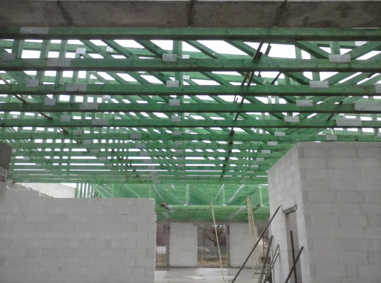 Wnętrze budynku w trakcie budowy, widoczne ściany z bloczków betonowych i zielona konstrukcja drewniana dachu z metalowymi łącznikami.